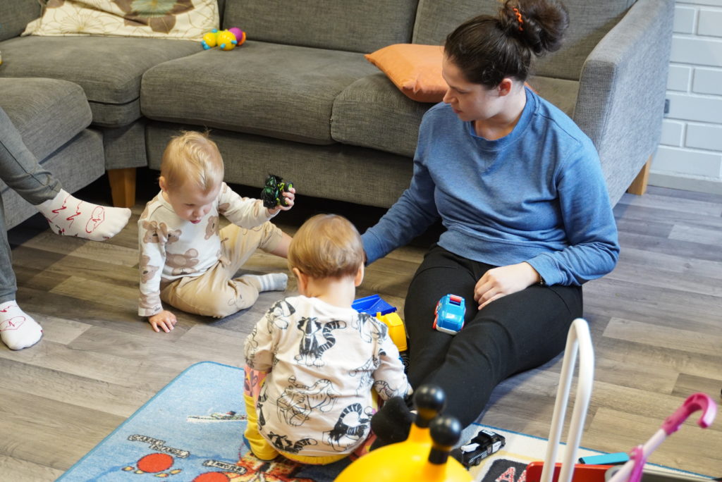 Lastenhoitaja istuu lattialla kahden taaperon kanssa ja leikkii heidän kanssaan.