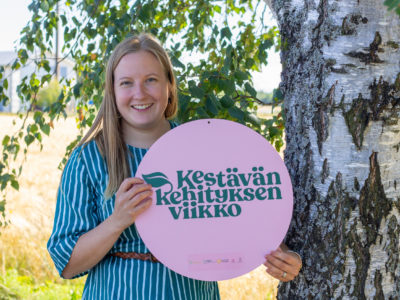 Nainen pitelee käsissään Kestävän kehityksen viikko -kylttiä aurinkoisena kesäpäivänä koivun alla.