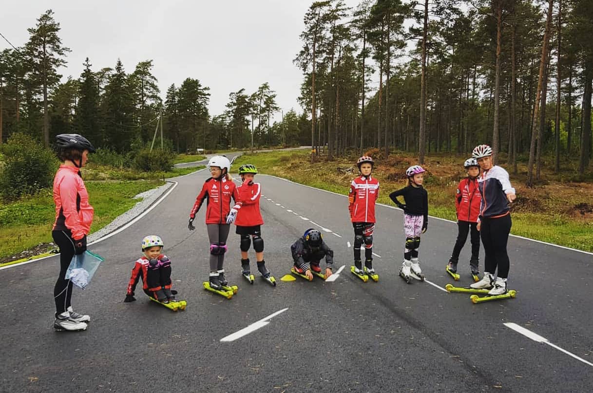 Keskellä seitsemän lasta rullasukset jalassa ja molemmissa reunoissa aikuinen neuvomassa.